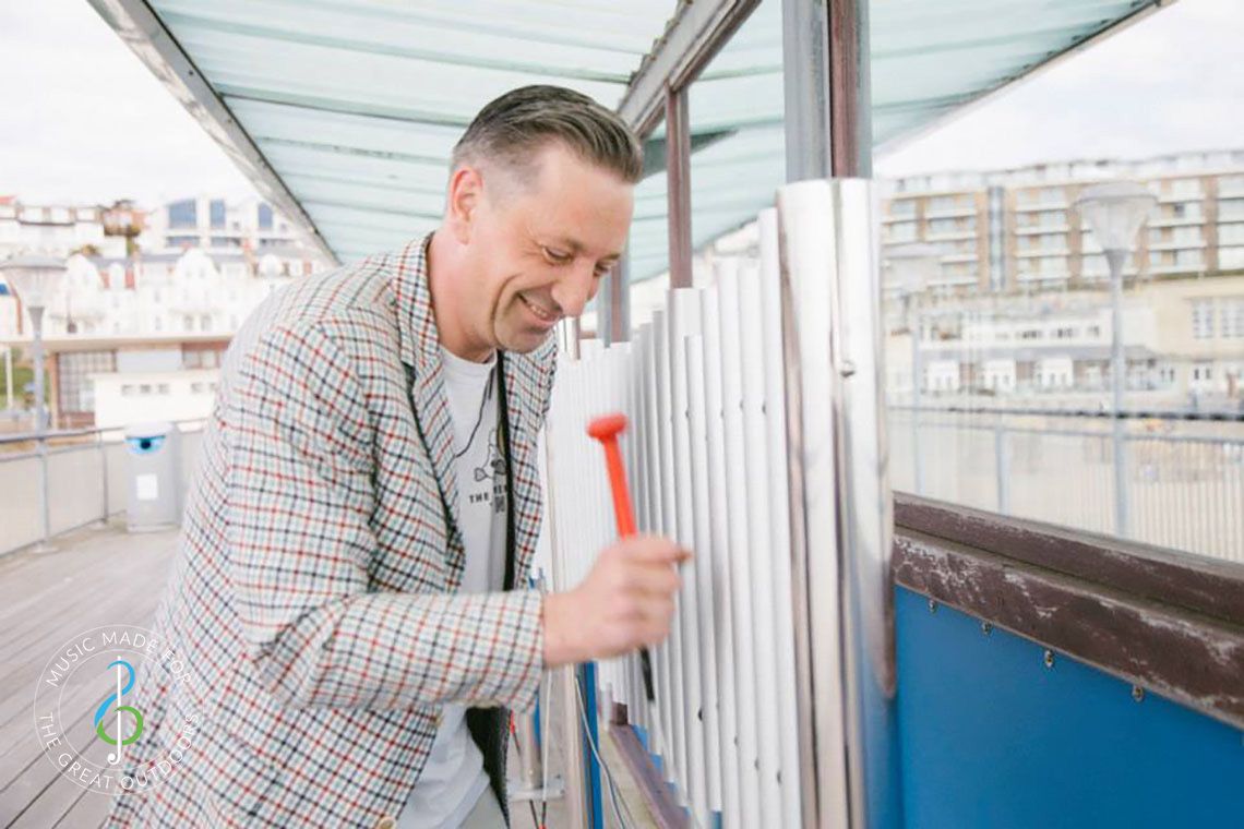 man hitting outdoor chimes with a red beater on a seaside pier