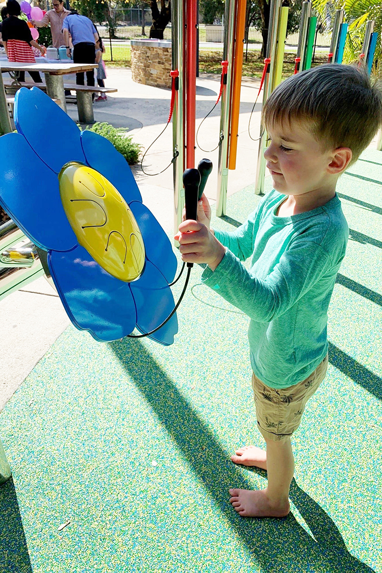 young barefoot boy playing a large outdoor musical drum shaped as a forget me not flower in a playground