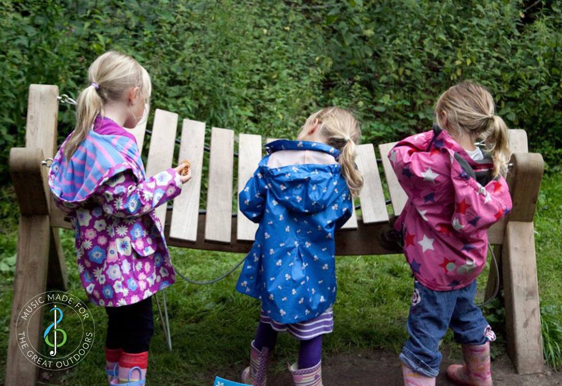 Three Young Girls in Raincoats Playing Large Outdoor Akadinda Xylophone