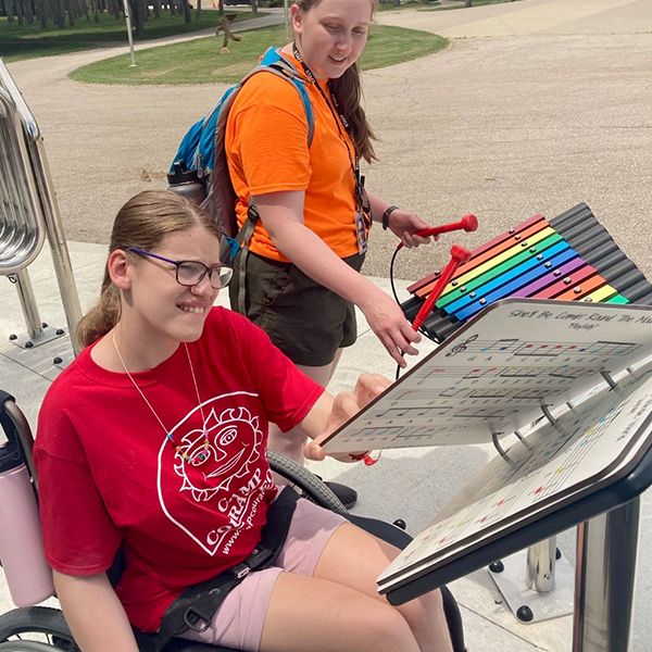 two teenage girls, one in a wheelchair, playing an outdoor rainbow colored xylophone in a music park at summer camp