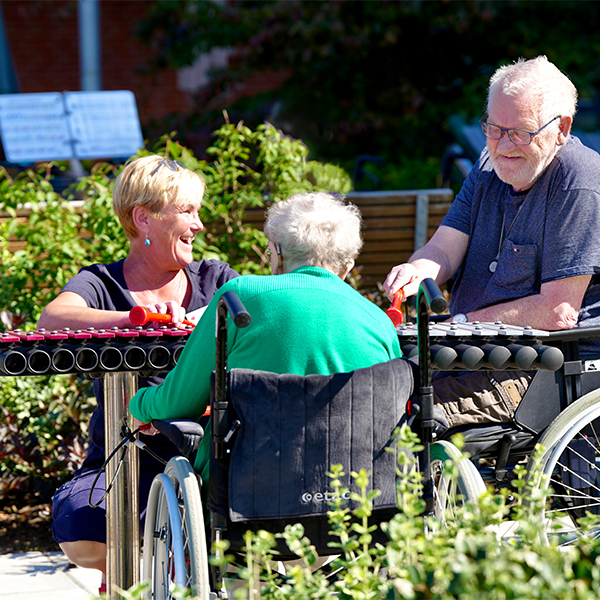 Two seniors in wheelchairs along with a carer playing on an outdoor xylophone in a memory garden