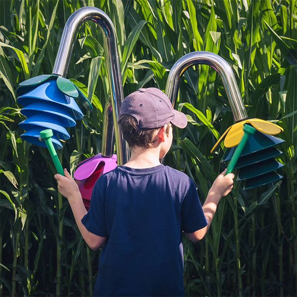 the back of a young boy striking a large outdoor musical instrument with two flower heads using a green mallet