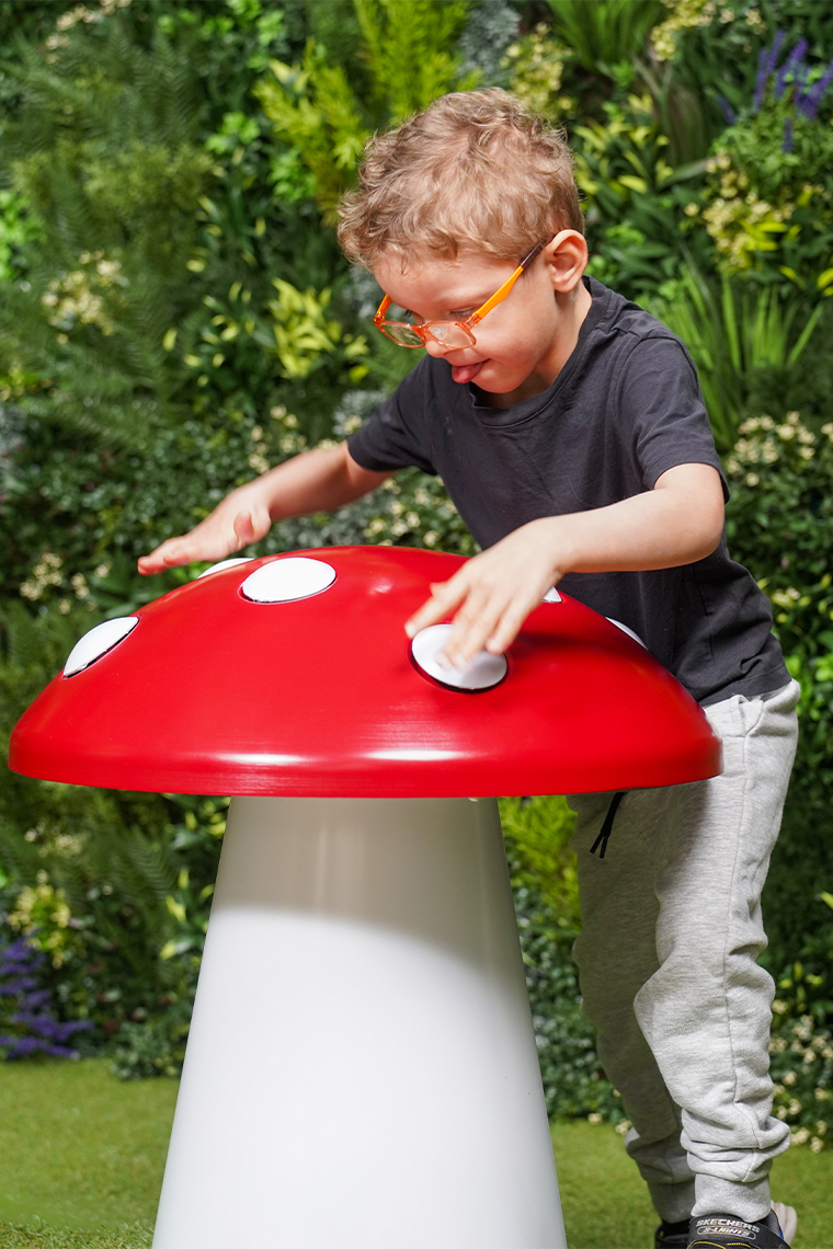 A young boy with glasses joyfully plays on a large red and white musical toadstool, surrounded by vibrant greenery, exuding a sense of wonder and playfulness.