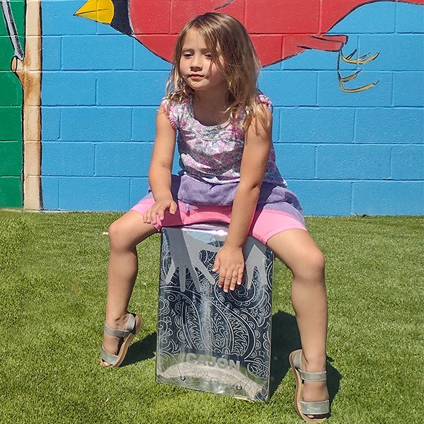 a young girl sitting on and playing a silver outdoor cajon drum at the town of versailles outdoor music garden