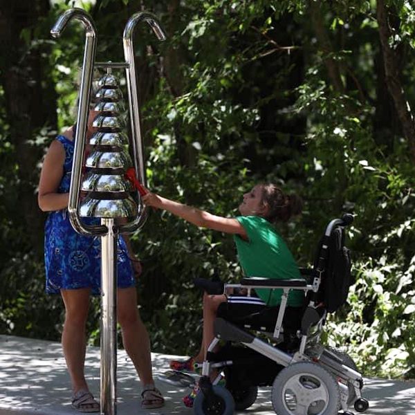 a young girl in a wheel chair playing a stainless steel bell lyre outdoor musical instrument in a summer camp playground