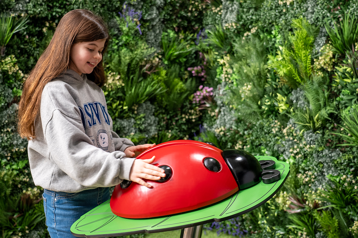 A girl in a gray hoodie playing a large musical ladybug on a green pedestal with her hands. The background is lush with green foliage, creating a playful, natural setting.