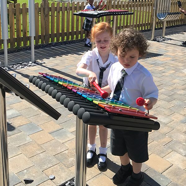 two young school pupils in school uniform playing a rainbwo colored outdoor xylophone