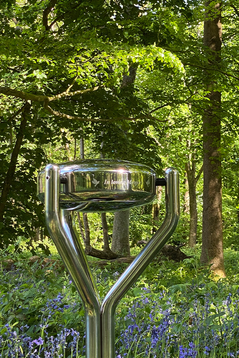 A large silver tongue drum engraved In Loving Memory in a forest setting surrounded by bluebells