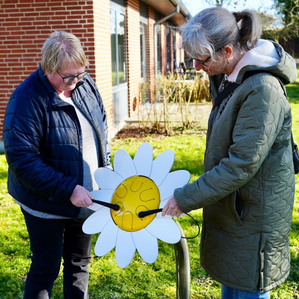 Two ladies playing an outdoor drum shaped like a daisy with white petals surrounding a yellow drum center in a care home sensory garden 