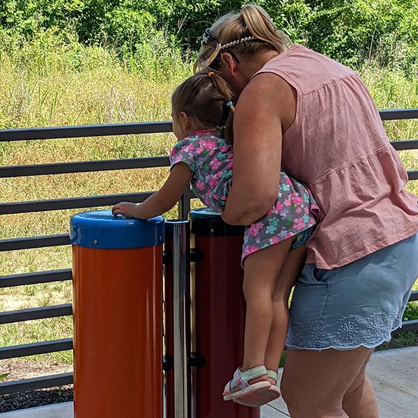 a young girl being help up by her mother to play colourful outdoor conga drums on the patio of the scenic regional library