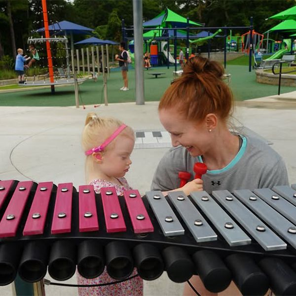 red haired lady and blond little girl behind a large outdoor xylophone with a play park in the background