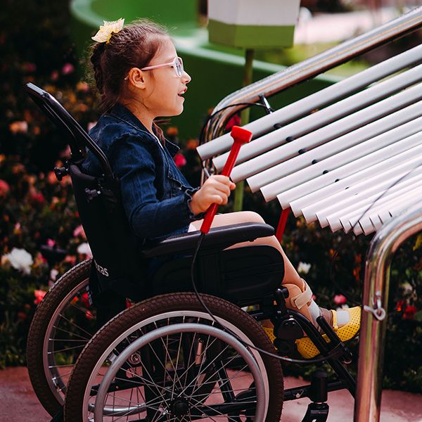 a young girl with special needs and in a wheelchair smiling and playing a large metal outdoor musical instrument in a playground