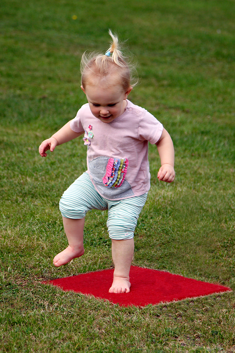 a young girl jumping on a red musical stepping stone square in a lawn which is disguising musical notes