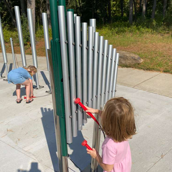 a young girl playing a large outdoor musical instrument in a musical playground in a park with another playing outdoor chimes in the background