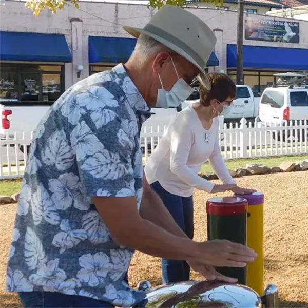 a man wearing a hat and a mask playing a stainless steel tongue drum at the sherman public library