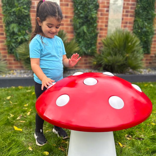 A child in a blue shirt smiles and touches a large, red mushroom-shaped musical sculpture with white spots. They are standing on green grass in a garden.