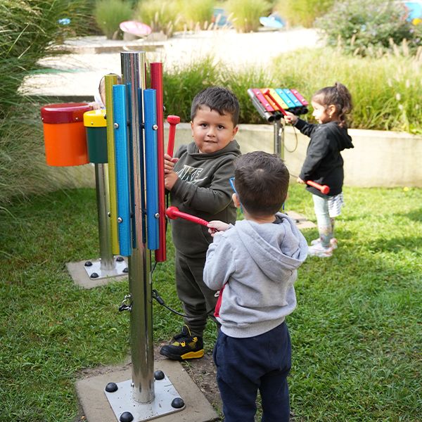 Tow young boys playing an outdoor musical chime with a small xylophone being played by a young girl in the background