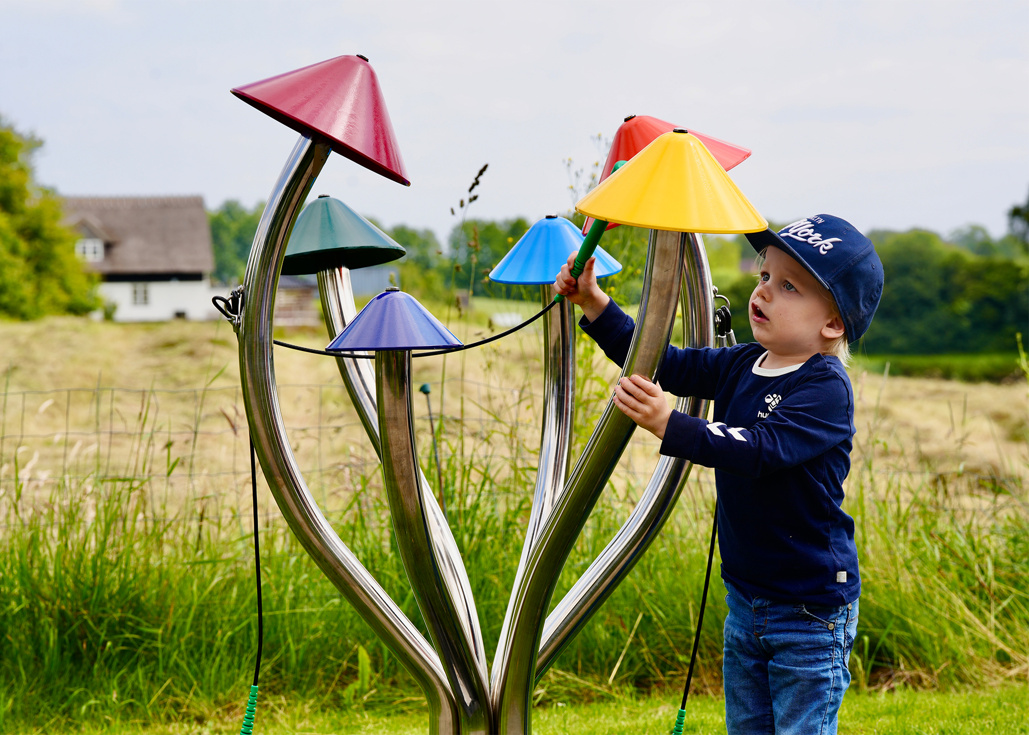 a young boy wearing a cap playing a large outdoor musical instrument shaped like field mushrooms with different coloured caps on each stalk