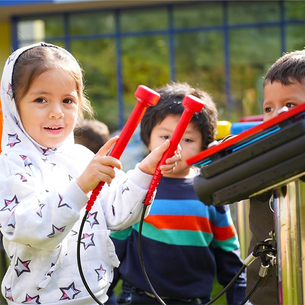 A young girl playing on a small outdoor xylophone being watched by other young children in a museum sensory garden