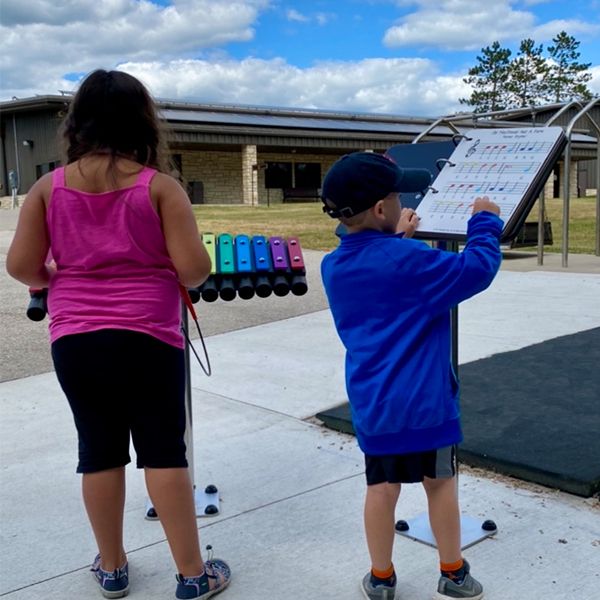 two children playing a large rainbow colored outdoor xylophone in a music park at summer camp