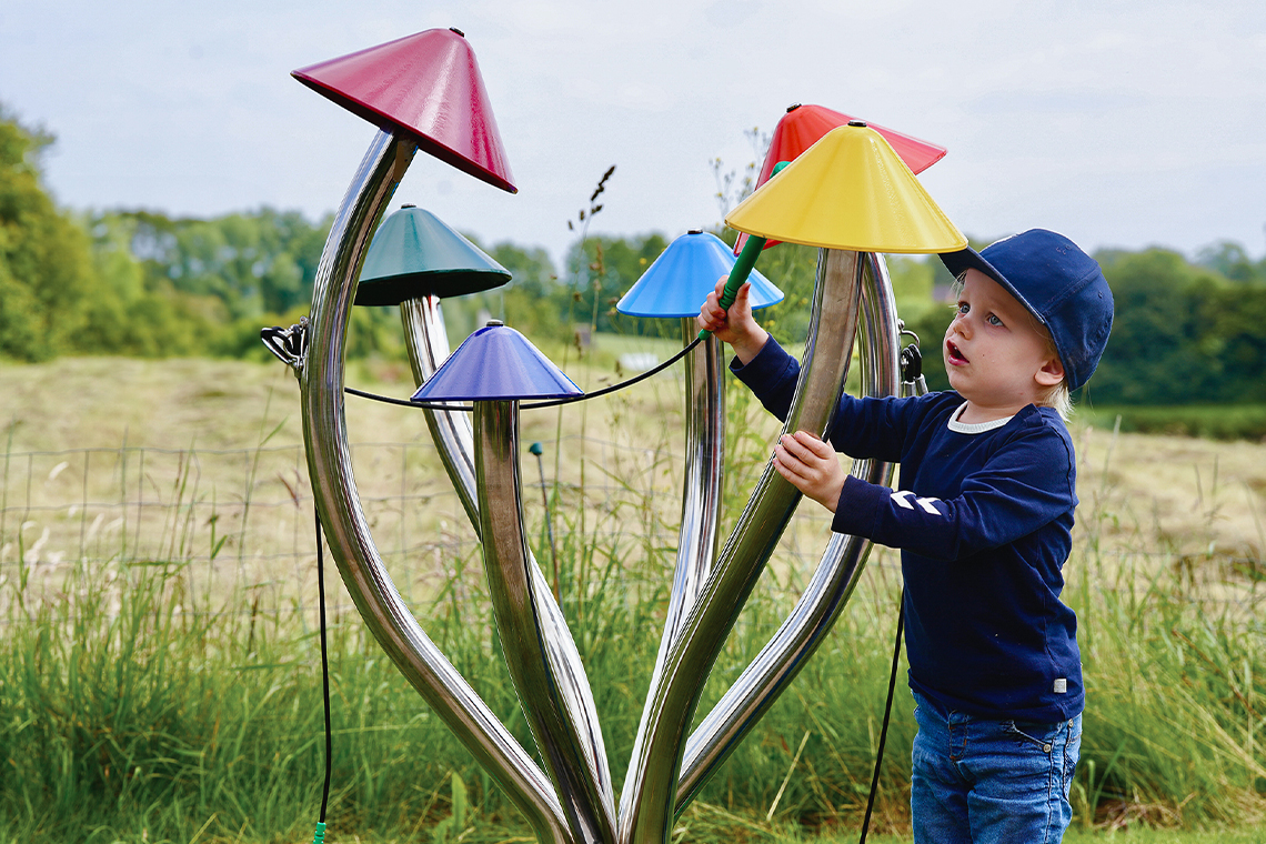 a young boy wearing a cap playing a large outdoor musical instrument shaped like field mushrooms with different coloured caps on each stalk