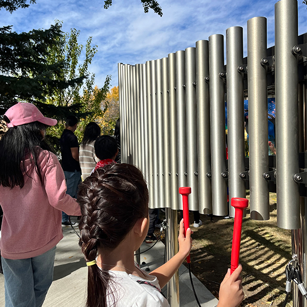 A child with braided hair plays an outdoor musical instrument, holding red mallets, while others watch in a park with autumn trees.