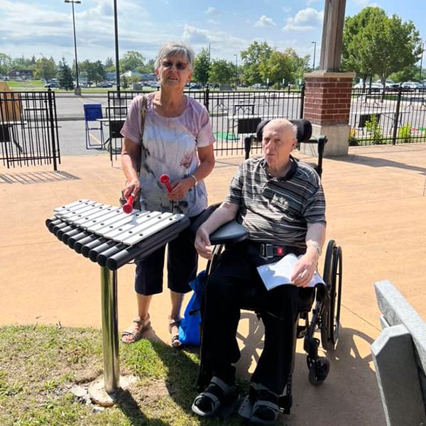 two seniors one standing and one in a wheelchair playing an outdoor musical instrument called a cadenza