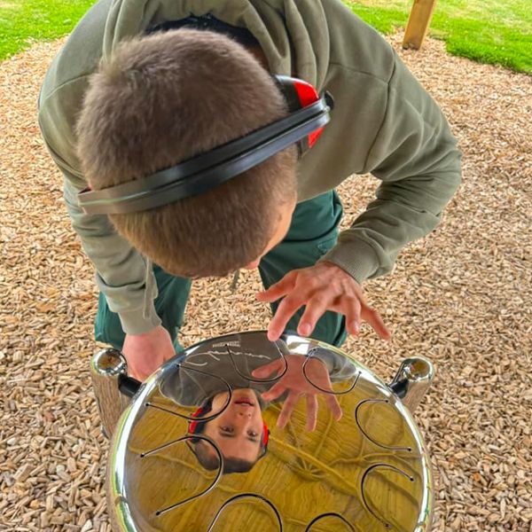 Young boy playing a shiny outdoor steel tongue drum, with his reflection visible on its surface at the Thomas Centre