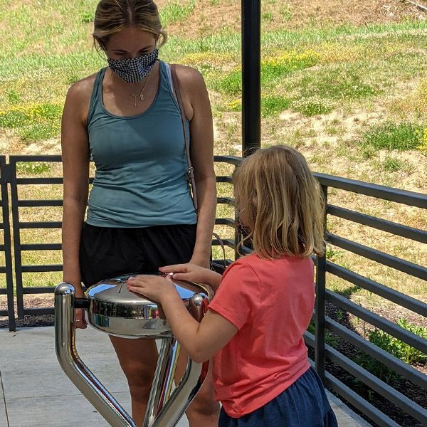 a young girl with blonde hair playing a stainless steel outdoor tongue drum with a female adult