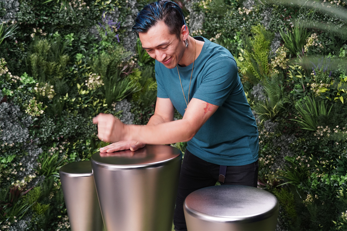 A person with blue hair smiles while playing sleek, metallic outdoor djembe drums in a lush, green garden setting, conveying joy and creativity.