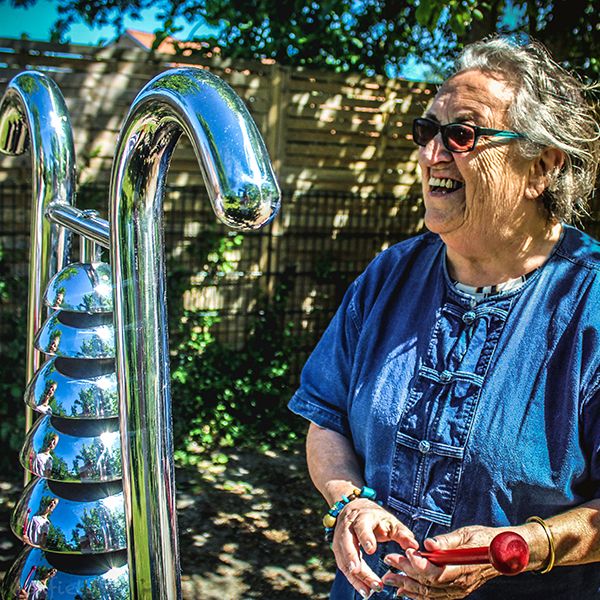 an older lady playing a large stainless steel outdoor musical instrument shaped like a bell lyre 