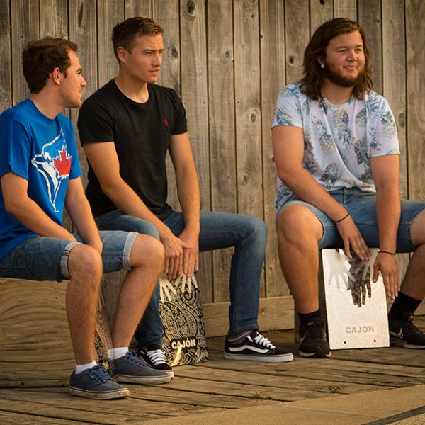 three young men sitting on stainless steel cajon percussion drums in an outdoor shelter