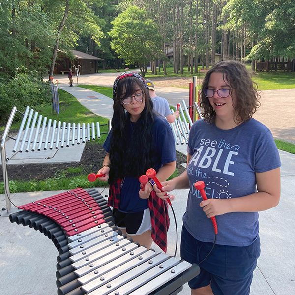 two female young adults with special needs playing a large outdoor xylophone in a music park at summer camp 