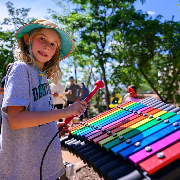 a young blond girl wearing a large rimmed hat playing a colourful outdoor xylophone