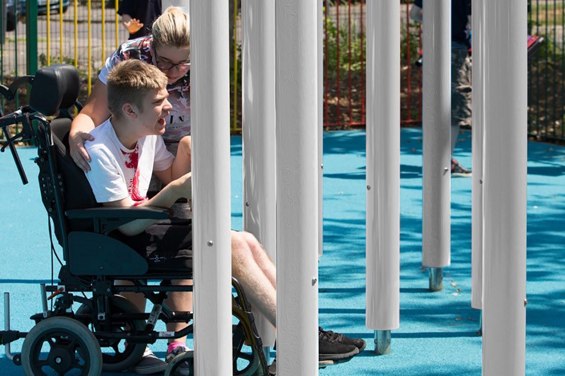 Young disabled man in a wheelchair with his carer playing on tall silver musical chimes in a school playground