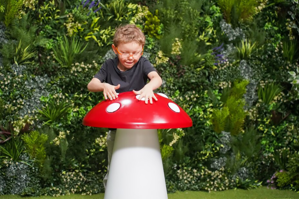 a young boy wearing glasses is playing a musical sculpture shaped like a toadstool, hitting the white dots