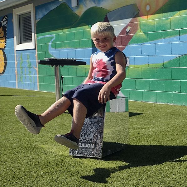 a young boy sitting on and playing a silver outdoor cajon drum at the town of versailles outdoor music garden