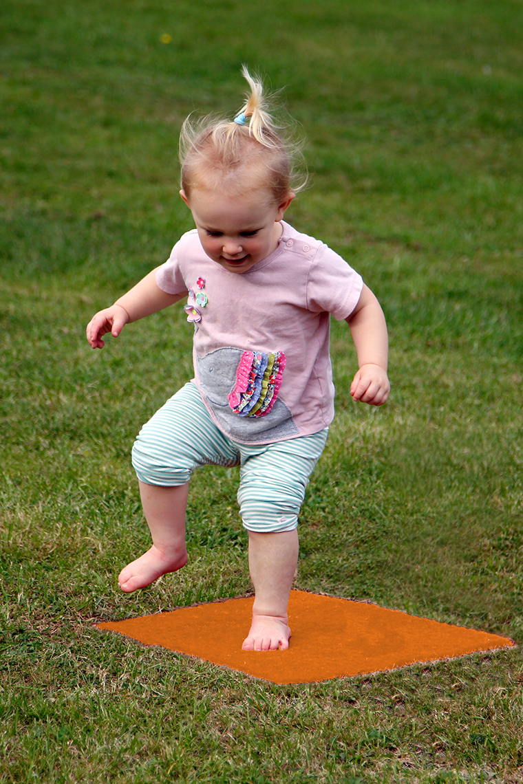 a young girl jumping on a orange musical stepping stone square in a lawn which is disguising musical notes