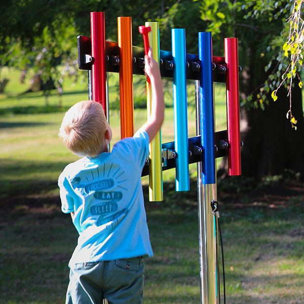 a young boy reaching up and playing on a set of six rainbow coloured outdoor musical chimes in a musical playground