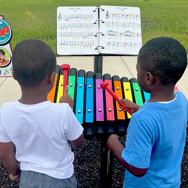 image of two young boys playing an outdoor xylophone with rainbow colored notes and a large outdoor music book in front