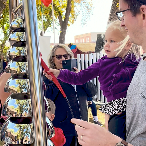 A father holds his young daughter as she taps shiny silver bell lyre with a mallet, surrounded by people at an outdoor event. They look engaged and curious.