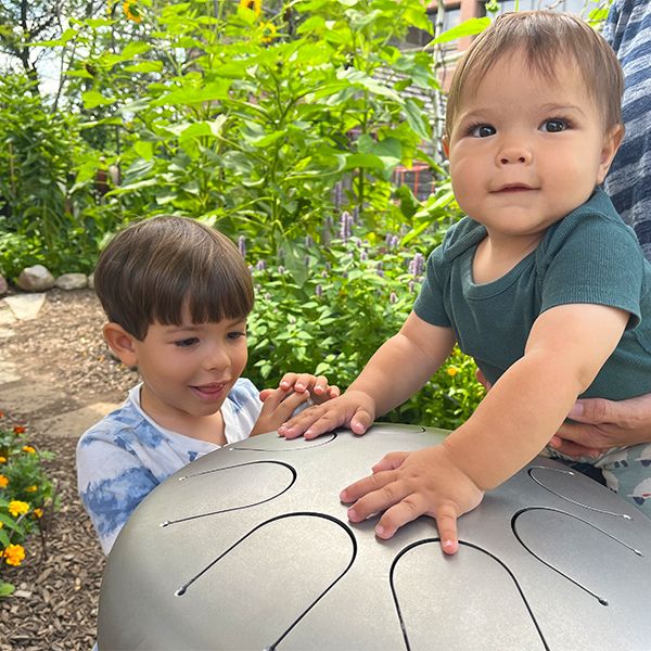 a young boy along with a baby being held up to play an outdoor stainless steel tongue drum in the Madison Childrens Museum