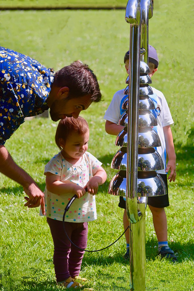 a very little girl and her father playing an outdoor stainless steel bell lyre musical instrument in a park