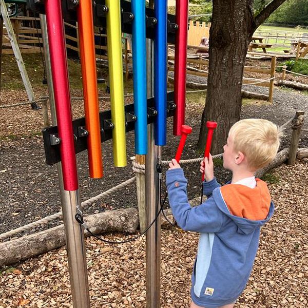 a young boy reaching up and playing a rainbow colored musical instrument in the bark filled musical playground at Bocketts Farm