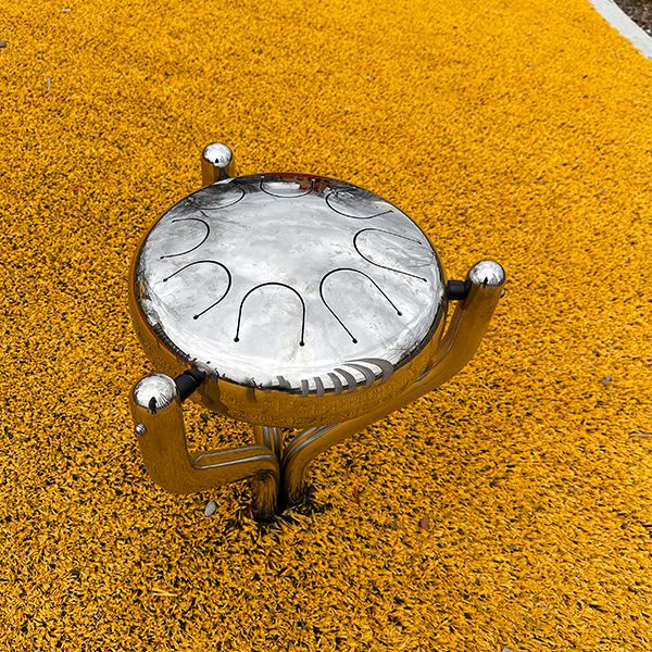 a large stainless steel outdoor tongue drum on yellow playground surfacing