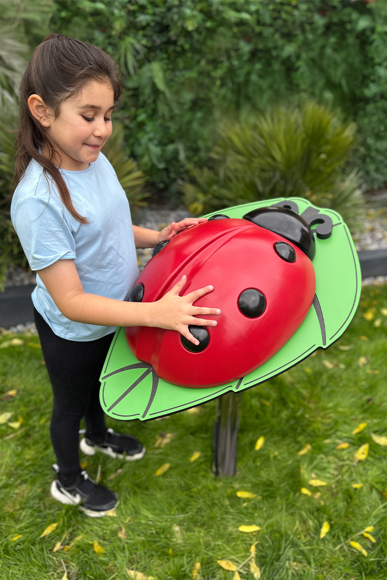 A young girl touching a large, red ladybug musical sculpture on a green leaf in a garden. She smiles with delight, surrounded by lush greenery.