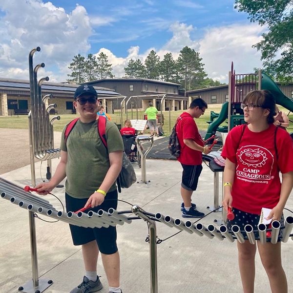 two young adults playing a large outdoor xylophone together in a music park at summer camp