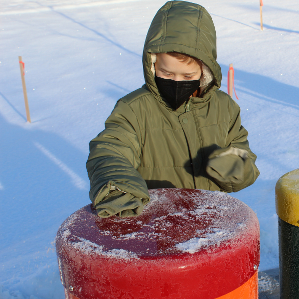 a young boy wearing a coat and a face mask playing outdoor musical drums at the clinton macomb public library