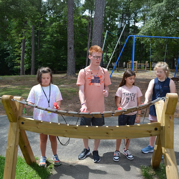 four children of varying heights playing a large wooden outdoor marimba in a camp ground