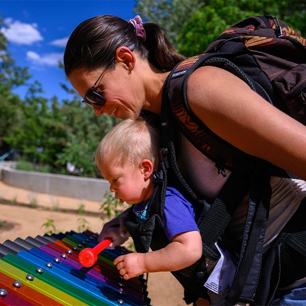 a mum with a baby in a carrier on her front bending over and playing a colourful outdoor xylophone in a music park